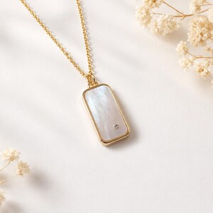 Può includere: Una collana a catena dorata con un ciondolo rettangolare caratterizzato da un inserto in madreperla bianca iridescente. Il ciondolo è incorniciato da un bordo in metallo color oro lucido e include un piccolo cristallo trasparente in basso a destra.