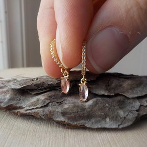 May include: Gold-toned hoop earrings with a delicate design. Each earring features a row of small, clear stones and a dangling, light pink, oval-shaped gemstone. The earrings are held against a textured, dark brown surface.