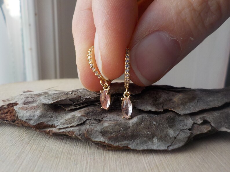 May include: Gold-toned hoop earrings with a delicate design. Each earring features a row of small, clear stones and a dangling, light pink, oval-shaped gemstone. The earrings are held against a textured, dark brown surface.