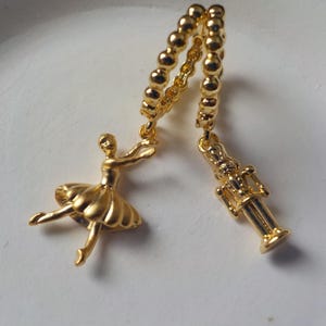 Può includere: Orecchini con ciondoli color oro raffiguranti una ballerina e uno schiaccianoci. La ballerina è in posa aggraziata e lo schiaccianoci è dettagliato. Gli orecchini sono collegati da una catena di piccole perle rotonde.
