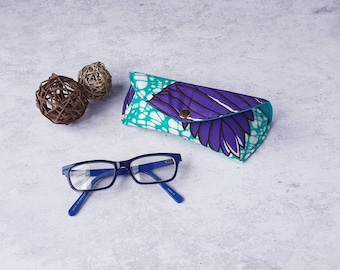 Op de afbeelding: Een blauw montuur bril ligt naast een brillenkoker met patroon. De koker heeft een turquoise en witte achtergrond met paarse veervormige ontwerpen. Twee decoratieve geweven bollen staan ook in de afbeelding.
