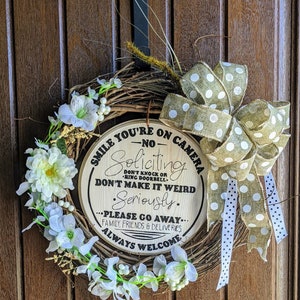 May include: A wooden wreath with a sign that reads "Smile you're on camera - No Soliciting. Don't knock or ring doorbell. Don't make it weird. Seriously. Please go away... Family, friends & deliveries. Always welcome". The wreath is decorated with white flowers and a brown and white polka dot bow.