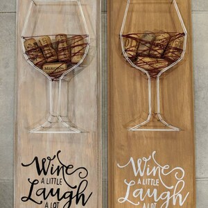 May include: Two wooden wine cork holders. Each features a wireframe wine glass design filled with corks, mounted on a wooden plaque. The phrase "Wine a little, laugh a lot" is written on each plaque.