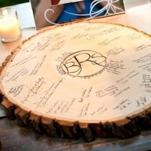 Puede incluir: Una rodaja de madera con un borde de corteza áspero, utilizada como libro de visitas en una boda. La superficie está cubierta de mensajes escritos a mano y las iniciales "B.R.S." están talladas en el centro.