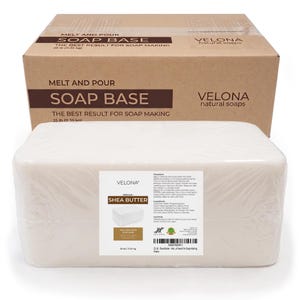 May include: A large rectangular block of white shea butter melt and pour soap base wrapped in clear plastic, positioned in front of a brown cardboard shipping box labeled Velona Natural Soaps, 25 lb (11.35 kg) soap base for soap making.