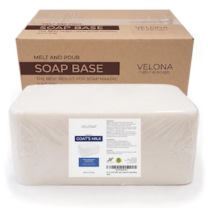 May include: A large brown cardboard box labeled Melt and Pour Soap Base 25 lb (11.35 kg) by Velona Natural Soaps, positioned behind a rectangular block of white goat's milk soap base wrapped in clear plastic with a detailed product label.