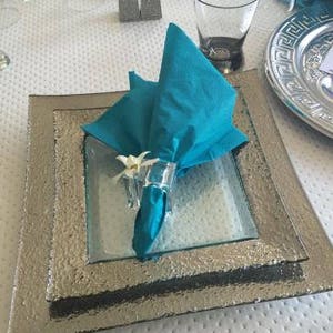 Pode incluir: Um guardanapo turquesa com uma flor branca &eacute; colocado em um prato quadrado prateado com um prato quadrado de vidro por baixo. O prato est&aacute; sobre uma toalha de mesa branca com um padr&atilde;o de pontos.