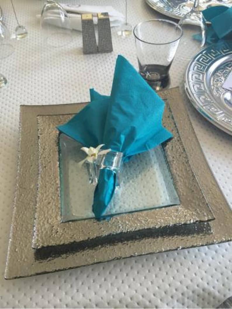 Pode incluir: Um guardanapo turquesa com uma flor branca &eacute; colocado em um prato quadrado prateado com um prato quadrado de vidro por baixo. O prato est&aacute; sobre uma toalha de mesa branca com um padr&atilde;o de pontos.