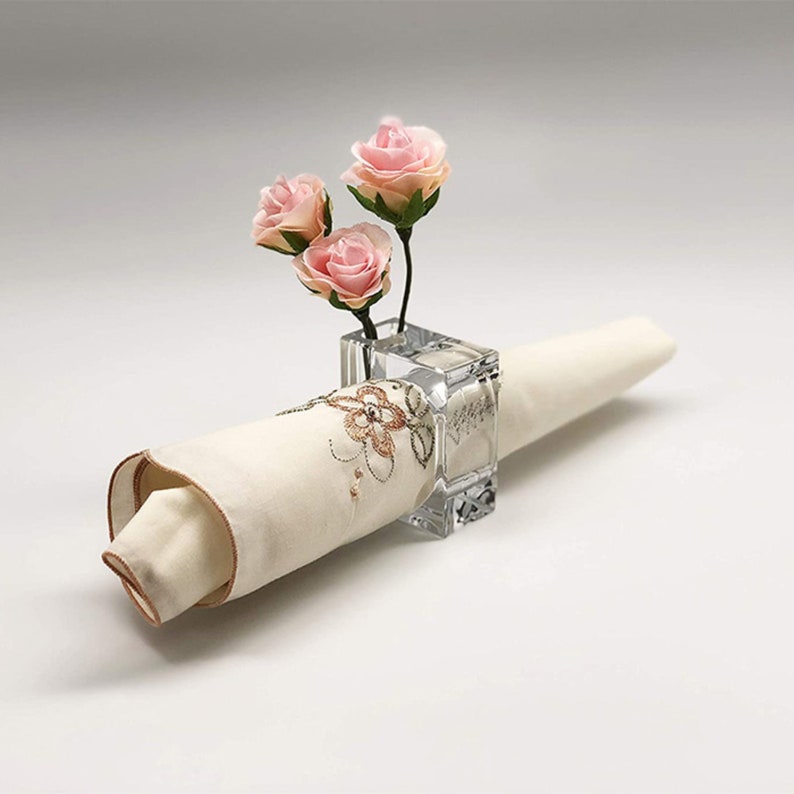 Pode incluir: Um anel de guardanapo de vidro transparente com um pequeno vaso no topo. O vaso cont&eacute;m tr&ecirc;s rosas de seda cor-de-rosa. O anel de guardanapo segura um guardanapo de linho branco com um desenho de bordado floral.