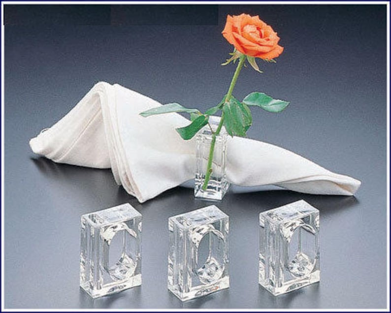 Pode incluir: Quatro argolas de guardanapo de acr&iacute;lico transparente com formato quadrado. As argolas s&atilde;o projetadas para segurar um guardanapo e um pequeno arranjo de flores. Uma &uacute;nica rosa laranja &eacute; exibida em um vaso de acr&iacute;lico transparente.