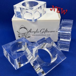 May include: Three clear acrylic napkin rings. The rings are square shaped with a round opening in the center. The rings are packaged in a white box with the words "Acrylic Giftware" and a website address printed on the box.