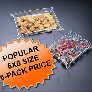 May include: Two clear plastic trays, one filled with small, round cookies and the other with colorful wrapped candies. The trays are square with a slightly raised edge. An orange and white sign reads "POPULAR 6X8 SIZE 6-PACK PRICE."