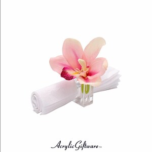 Pode incluir: Um porta-guardanapo de acr&iacute;lico transparente com uma flor de orqu&iacute;dea rosa. O porta-guardanapo &eacute; um design simples com uma base quadrada e um cilindro transparente.