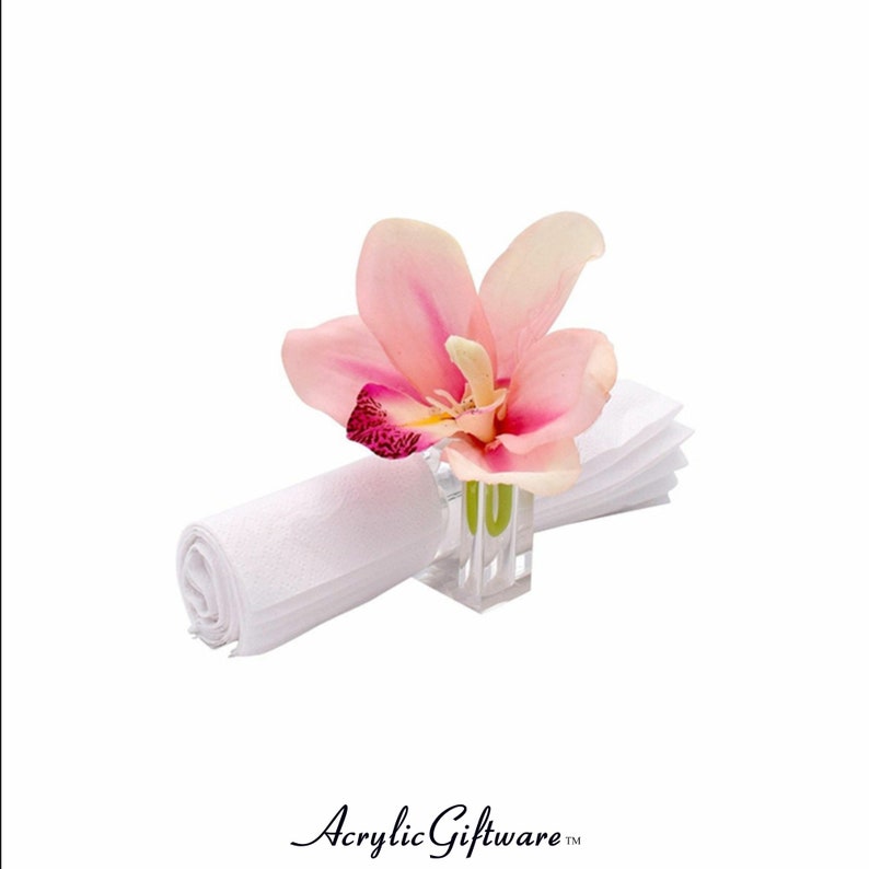 Pode incluir: Um porta-guardanapo de acr&iacute;lico transparente com uma flor de orqu&iacute;dea rosa. O porta-guardanapo &eacute; um design simples com uma base quadrada e um cilindro transparente.