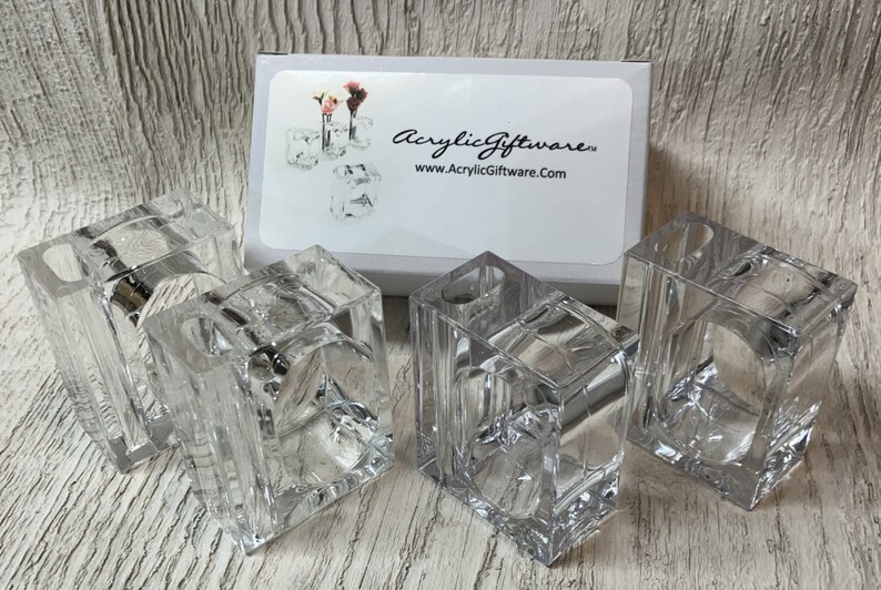 Pode incluir: Quatro argolas de guardanapo de acr&iacute;lico transparente, cada uma com formato quadrado e uma abertura ligeiramente arredondada. As argolas est&atilde;o dispostas sobre uma superf&iacute;cie de madeira clara. As argolas s&atilde;o feitas pela AcrylicGiftware, como indicado na caixa branca no fundo.