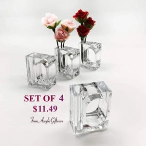 May include: Set of four clear acrylic napkin rings with a small vase for a single flower. The rings are square shaped and have a smooth finish.  The text "SET OF 4 $11.49 From Acrylic Giftware" is visible in the image.