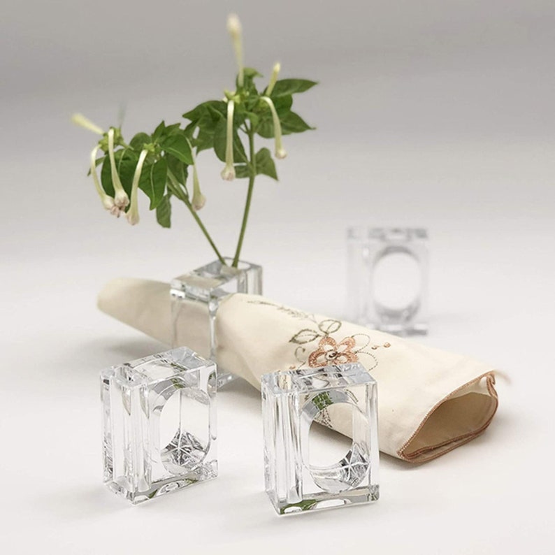 Pode incluir: Quatro argolas de guardanapo de acr&iacute;lico transparente. As argolas s&atilde;o quadradas com uma abertura arredondada. As argolas est&atilde;o sobre um guardanapo de linho branco com um padr&atilde;o floral. Um pequeno buqu&ecirc; de flores brancas est&aacute; no fundo.