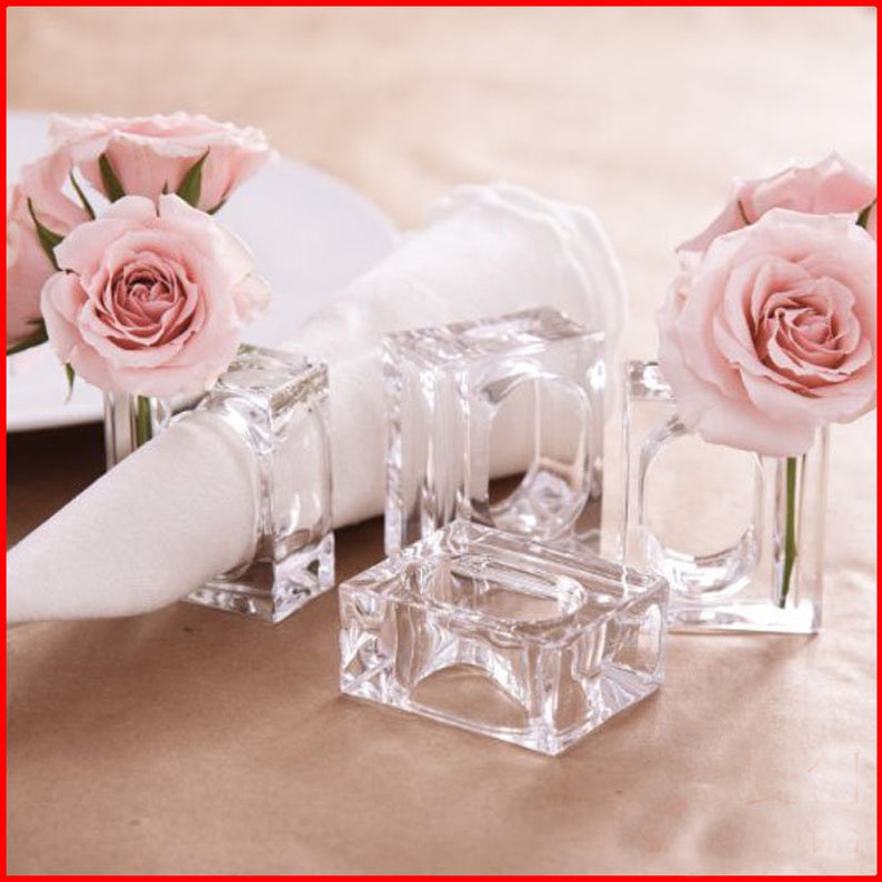 Pode incluir: Quatro argolas de guardanapo de vidro transparente com um design quadrado. Cada anel tem uma pequena abertura na parte superior para segurar uma &uacute;nica rosa rosa.
