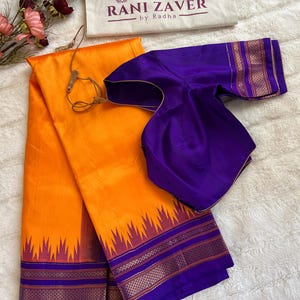 May include: An orange saree with a purple and gold border, paired with a purple blouse. The saree has a decorative border with a zig-zag pattern. The brand name "RANI ZAVER by Radha" is visible on a cream-colored bag.