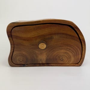 May include: A handcrafted wooden box with a unique, organic shape. The box is made of dark brown wood, featuring a lid with a small, round wooden knob. The wood grain is visible, adding to the natural aesthetic. The box is designed with a curved edge.