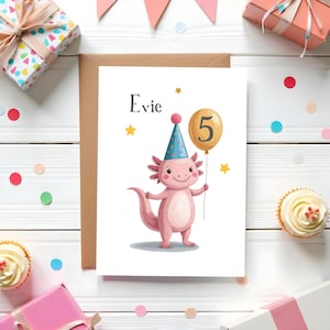 Peut inclure: Carte d'anniversaire avec un axolotl rose portant un chapeau de fête et tenant un ballon numéro 5. La carte porte le nom "Evie" en haut, avec des étoiles et des confettis. Des cadeaux et des cupcakes entourent la carte.