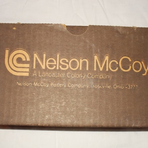 May include: A brown cardboard box with the text "Nelson McCoy, A Lancaster Colony Company, Nelson McCoy Pottery Company, Roseville, Ohio -3777" printed in yellow.