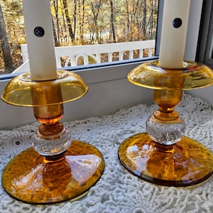 May include: Two amber glass candle holders with clear glass bases and a textured bubble design. The candle holders are on a white crocheted surface.