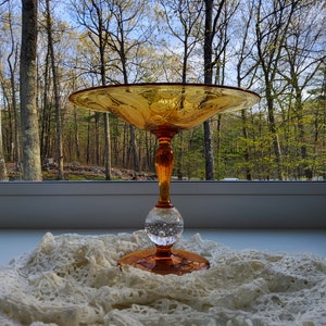 May include: A vintage amber glass pedestal bowl with a clear glass ball base. The bowl has a scalloped edge and intricate etched designs. It is sitting on a white lace tablecloth.