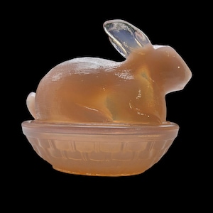 May include: A vintage glass rabbit butter dish in a pale pink color. The rabbit is sitting in a basket-shaped base.