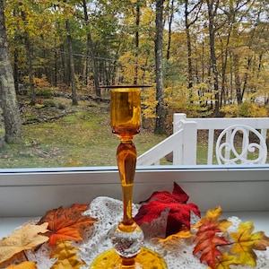 May include: Amber glass candle holder with a clear glass ball at the base. The holder is on a white crocheted doily with fall leaves.