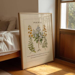 May include: Framed botanical print featuring a central illustration of a flowering plant with white and yellow blooms. The text "PSALM 1:3" is at the top, with a Bible verse below. The frame is light-colored, and the print is leaning against a wall.