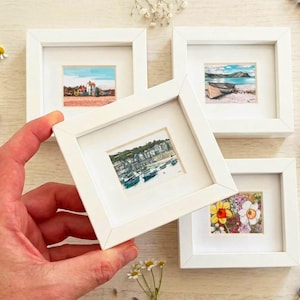 May include: Four small square white frames containing miniature landscape and floral paintings. One frame is held in the foreground, displaying a harbour scene with boats, while the others show a house, a beach with a boat, and colourful flowers.
