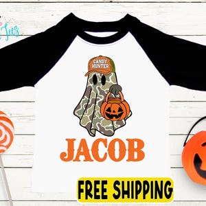 May include: White and black raglan-style Halloween shirt with a ghost graphic wearing a camo hat and holding a pumpkin bucket. The name "JACOB" is printed in orange below the graphic. A lollipop and pumpkin bucket are also in the image.