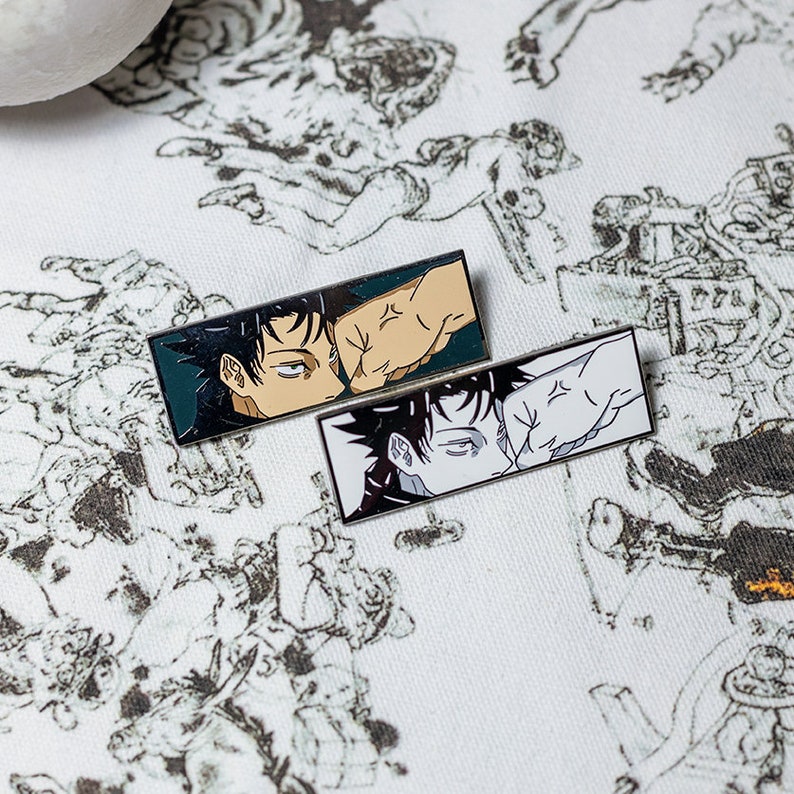 May include: Two rectangular enamel pins featuring anime-style illustrations. One pin has a black and tan color scheme, while the other is black and white. Both pins depict a fist making contact with a face, with a teal and white background.