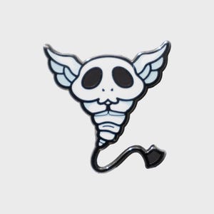 May include: A white and black enamel pin featuring a stylized skull with wings and a tail. The skull has large black eye sockets and a small mouth. The wings are white with light blue accents. The tail ends in a black spade shape.
