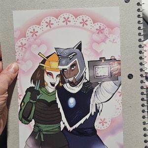 May include: A print of two characters in cosplay, one in green and black armor, the other in blue armor, posing for a selfie. The background has a pink floral and heart design.