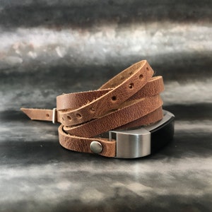 Puede incluir: Una banda de cuero marrón para un rastreador de actividad física. La banda está envuelta alrededor del rastreador y asegurada con un cierre de metal plateado.