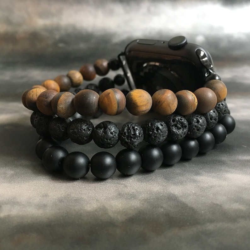 May include: Three layered beaded bracelets in black, brown, and black. The top layer is brown tiger's eye beads, the middle layer is black lava rock beads, and the bottom layer is matte black beads. The bracelets are worn on a black Apple Watch.