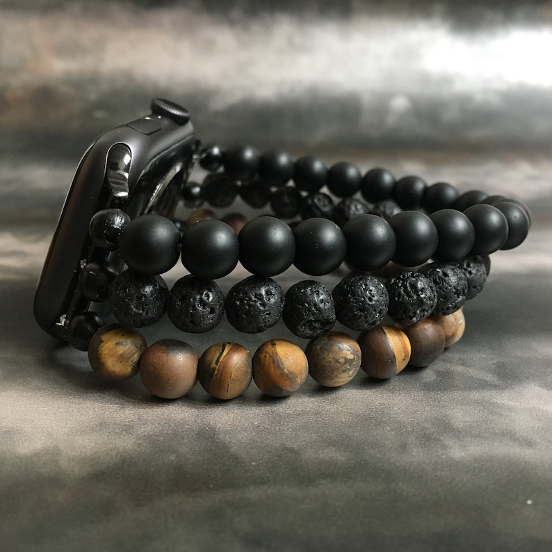 May include: Three beaded bracelets stacked on a black Apple Watch. The top bracelet is black, the middle bracelet is black lava rock, and the bottom bracelet is brown tiger's eye.