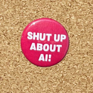 May include: A circular pinback button with a vibrant magenta background and bold white text that reads SHUT UP ABOUT AI! The button has a smooth, glossy finish and a simple, clean design that stands out clearly against the textured surface.
