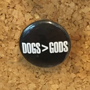 May include: A black button with white text that reads "DOGS>GODS".