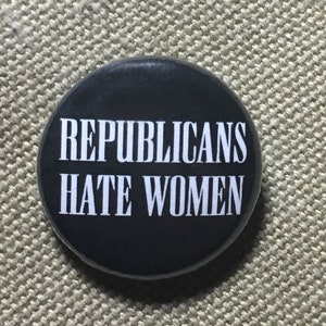 May include: A round, black button with the white text "REPUBLICANS HATE WOMEN". The button is centered and slightly angled, set against a textured, tan background.