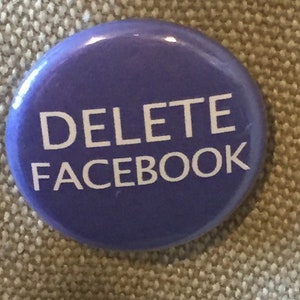 May include: A round, purple button with the words "DELETE FACEBOOK" in white, sans-serif font. The button has a glossy finish and is set against a neutral, textured background.