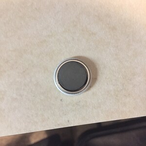Puede incluir: Un im&aacute;n redondo de color plateado con un centro negro.