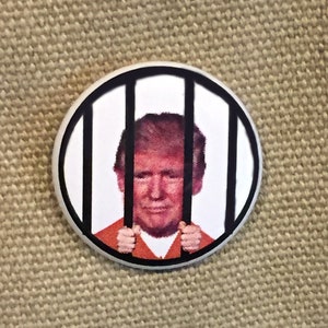May include: A white button with a black border depicting a person with orange hair and a orange jumpsuit behind black bars.