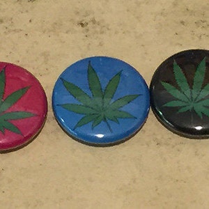 May include: Five round buttons in a row, each with a green cannabis leaf design. The buttons are yellow, pink, blue, black, and red. The buttons are on a light brown surface.