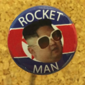 May include: A blue and red button with the text "ROCKET MAN" in white. The button features a photo of a man with dark hair wearing sunglasses.