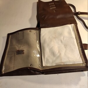 Puede incluir: Una bolsa de cuero marr&oacute;n con una solapa y un bolsillo transparente de pl&aacute;stico. La bolsa tiene una correa y un forro blanco.