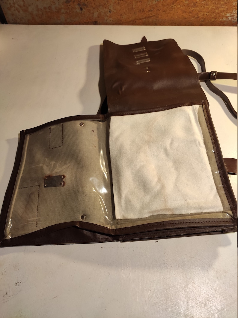 Puede incluir: Una bolsa de cuero marr&oacute;n con una solapa y un bolsillo transparente de pl&aacute;stico. La bolsa tiene una correa y un forro blanco.