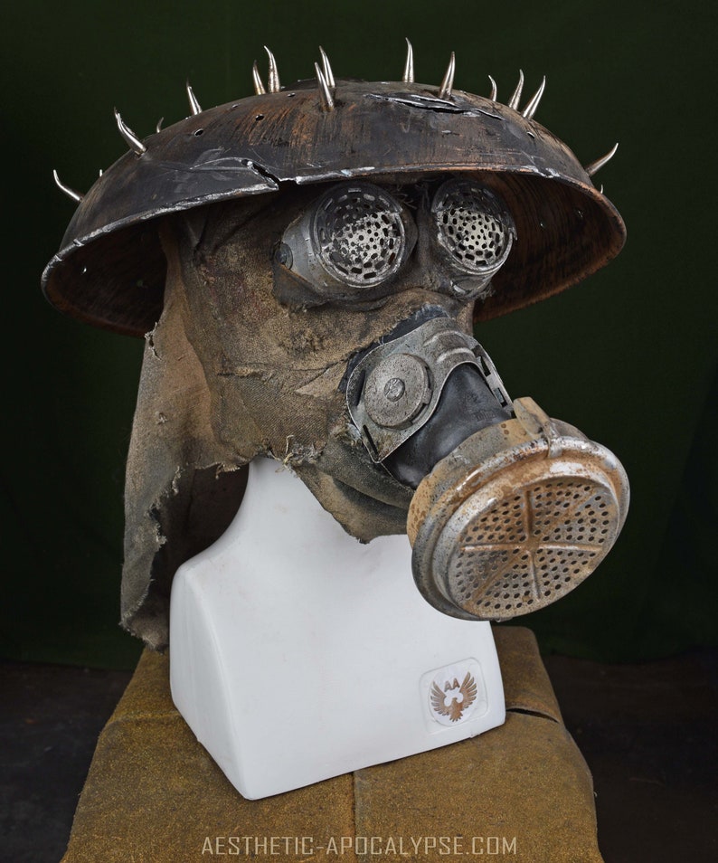 May include: A weathered, brown gas mask with a metal helmet and spikes. The mask has two round, black eye pieces and a large, round filter. The helmet is covered in dirt and grime.